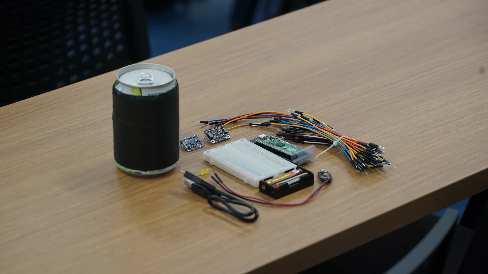 机の上に並べられた缶サットの材料一式。黒いテープを巻いた空き缶、ブレッドボード、ジャンパワイヤ、センサモジュール、Raspberry Pi Pico、電池ボックス